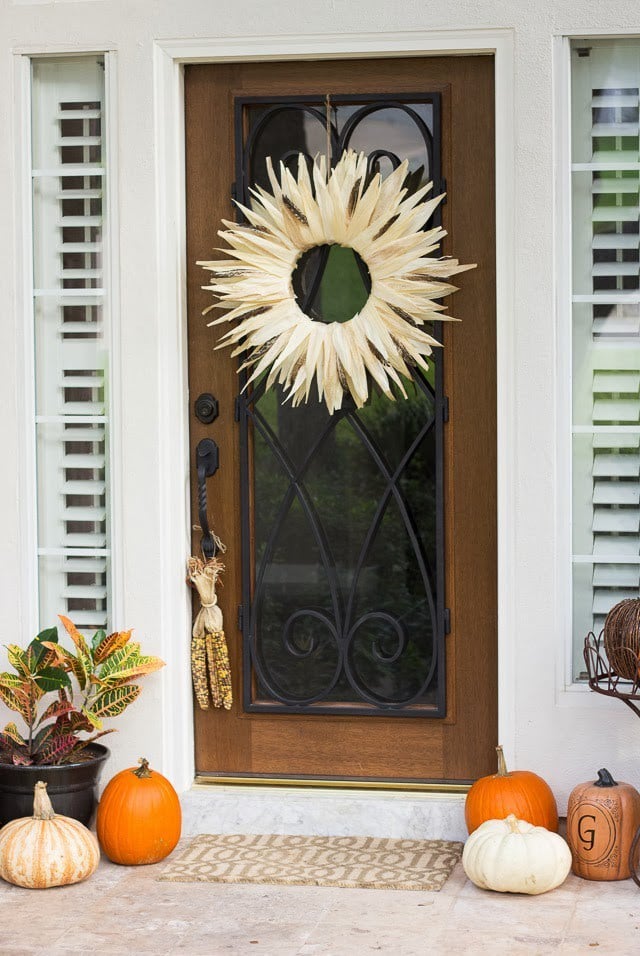 Corn Husk Wreath In Circle With Open Center On A Wooden Door With Glass Center And Fall Porch With Pumpkins And Hanging Corn Cobs